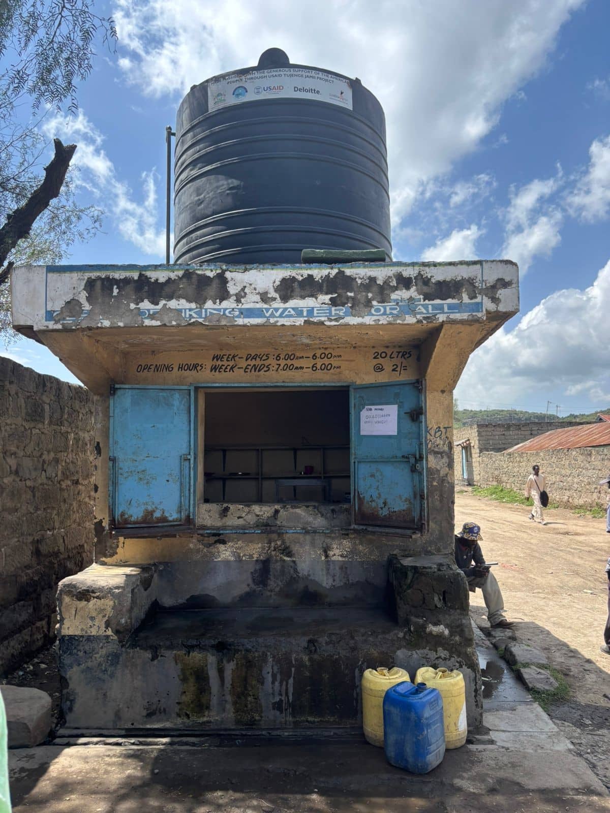 Waterkiosk met watertank op het dak en jerrycans ervoor. De dagelijkse openingstijden staan boven de ingang.
